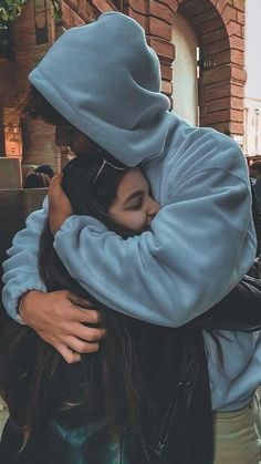 two people hugging each other in front of a brick building on a city street, one is wearing a blue hoodie and the other has her hair pulled back