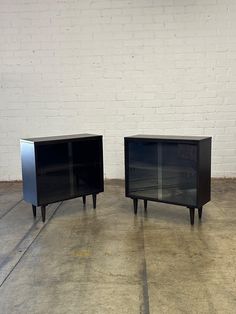 two black cabinets sitting next to each other on top of cement floored ground in front of a white brick wall