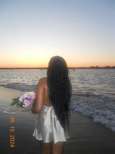 a woman standing on the beach with her back to the camera and flowers in her hand