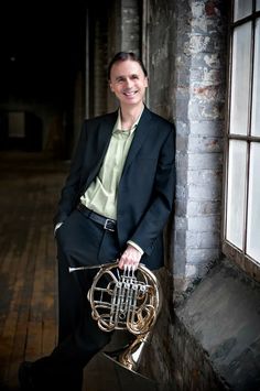 a man in a suit and tie holding a french horn while leaning against a brick wall