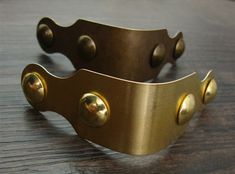 two brass bracelets on a wooden table with metal rivets and holes in the middle