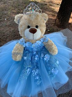 a teddy bear wearing a blue dress and a tiara sitting on the ground next to a tree