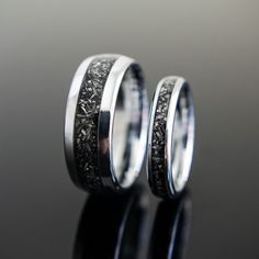 two wedding bands with black diamonds in them on top of a reflective surface, one is white gold and the other is silver