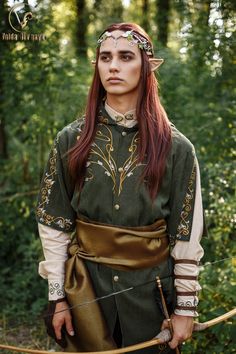 a woman with red hair is dressed in medieval clothing and holding a bow while standing in the woods