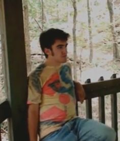 a young man sitting on top of a wooden porch next to a forest filled with trees