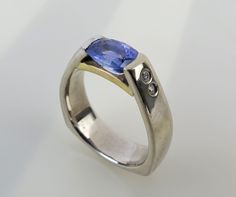 "A fancy faceted Tanzanite tops this wonderful minimalist 14k white & yellow gold diamond accent ring. The 7mm x 5.4mm x 3.7mm lavender blue oval faceted gem is tension-set tightly between thick solid bands of white gold. A yellow gold bridge supports the gem below. Two diamonds (1mm and 1.2mm) are bezel-set on each shoulder (.028 carat total). The ring was finished with a Euro or European shank, which means it has a flattened bottom that sits on the underside of the finger. Generally this style Rhodolite Garnet Ring, Diamond Accent Ring, Yellow Gold Diamond Ring, Faceted Gems, Blue Tanzanite, Gold Diamond Ring, Purple Band, Lavender Blue, Round Rings