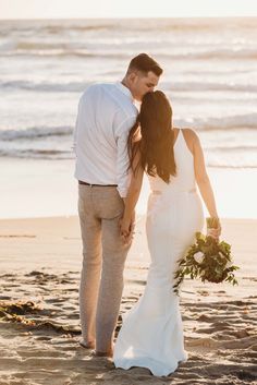 a bride and groom walking on the beach at sunset with their back to the camera