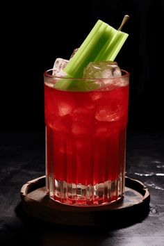 a red drink with celery sticks in it on a wooden tray next to a black background