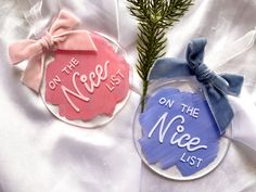 two ornament ornaments with bows and ribbons on white satin material, one has the words on the nice list