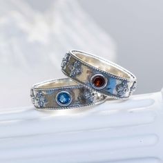 two silver rings with blue and red stones on them sitting on a white tablecloth