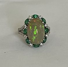 Ring in white gold (18k) centered with an oval cabochon opal of approximately 5.24 ct (16.15 x 11 mm) in an alternating surround of shuttle-cut "vivid green" emeralds and round brilliant-cut diamonds. Frame partially paved with diamonds. Total weight of emeralds: approximately 0.82 ct. Total weight of diamonds: approximately 0.41 ct. GFCO certificate for the 3 stones. Eagle's head hallmark and Jeweler's hallmark. Top height: 11 mm. Gross Weight: 5.81 g. Size: 54 Possibility of sizing, contact us Oval Multi-stone Emerald Ring, Fine Jewelry Oval Cabochon Opal Ring, Oval Cabochon Opal Ring Fine Jewelry, Oval Cabochon Opal Ring In Fine Jewelry, Formal Oval Cabochon Opal Ring, Elegant Green Opal Ring Oval Cabochon, Elegant Green Cabochon Opal Ring, Elegant Green Opal Ring With Oval Cabochon, Elegant Green Opal Oval Ring