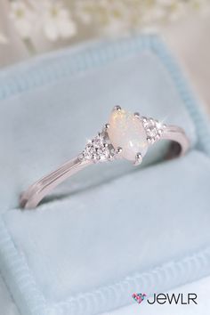 a white opal and diamond ring sitting on top of a blue velvet box with flowers in the background