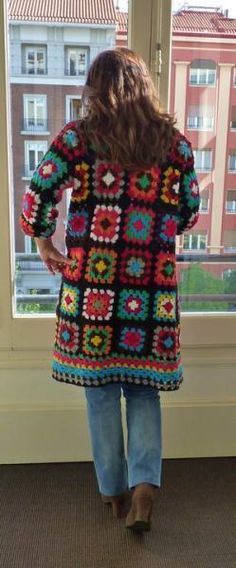 a woman standing in front of a window wearing a multicolored crocheted jacket