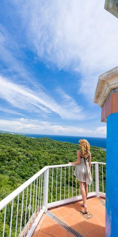 a woman standing on top of a balcony next to a lush green forest under a blue sky