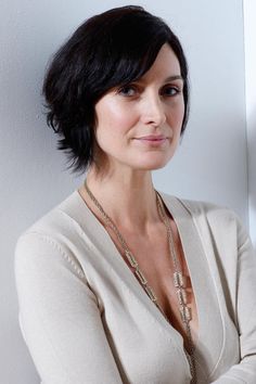 a woman with her arms crossed wearing a white shirt and gold necklace, leaning against a wall