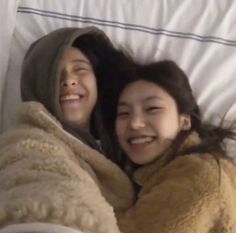 two young women laying in bed under a blanket and smiling at the camera with their arms wrapped around each other