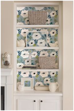 a white book shelf with baskets and flowers on it in front of a blue wallpaper