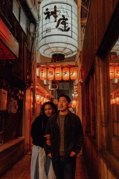 two people standing in an alley with lanterns on the ceiling and lights hanging above them