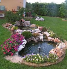 a small pond in the middle of a lawn with flowers and rocks around it, next to a house