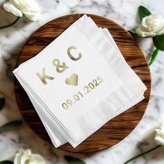 two napkins with gold foil on them sitting on top of a wooden plate next to white flowers