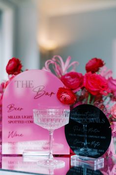 a table topped with pink flowers next to a glass filled with liquid and a sign that says the bar