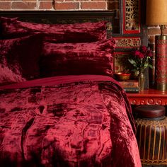 a bed with red comforter and pillows in front of a brick wall next to a lamp