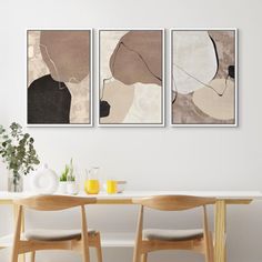 three abstract paintings hang on the wall above a dining room table with two chairs and a vase