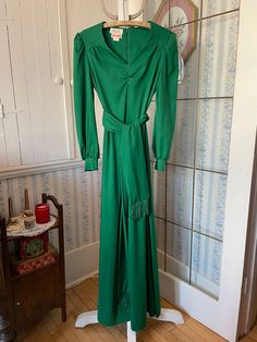 This stunning dress is made from medium weight polyester knit in rich emerald green. It has slightly puffy sleeves, gathers on the yoke and sleeves, cloth covered buttons on the cuffs, and a matching belt or scarf trimmed with silky emerald green fringe. The dress fastens in the back with a hook and eye closure and long zipper. Marked sized 13. The measurements, taken with the dress lying flat, are: shoulder to shoulder, 15 inches; armpit to armpit, 18 inches; waist, 17 inches; sleeves, 24 inches; length, 57 inches; bottom edge, 50 inches. The belt measures 75 inches long by 5 1/2 inches wide, with 3 1/2 inch fringe on each side. The fabric has quite a bit of stretch. In very good condition. Vintage Green Maxi Dress For Evening, Green Long Sleeve Maxi Dress For Fall, Green Maxi Dress For Fall, Formal Green Maxi Dress For Fall, Green Formal Maxi Dress, Vintage Long Green Dress, Green Belted Evening Dress, Green Maxi Dress For Evening, Vintage Green Maxi Dress For Party