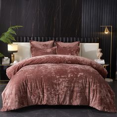 a bed with a pink comforter and pillows on it in front of a black wall