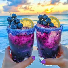 two people holding up glasses with blueberries and lemons on the beach at sunset