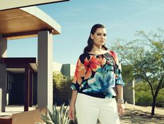 a woman standing in front of a house wearing white pants and a colorful top with flowers on it