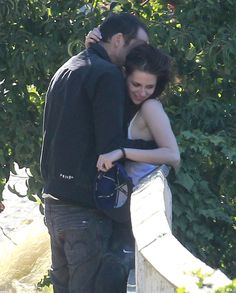 a man and woman embracing each other in front of some bushes with trees behind them