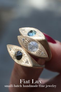 A thumb and index finger hold three yellow gold rings in the shape of an eye, set with a round inverted brilliant cut black diamond, salt and pepper diamond, and blue Montana sapphire. Cartier Panthere, The Third Eye, Diamond Eyes, Salt And Pepper Diamond, Eye Jewelry, Pretty Jewellery