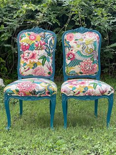 two blue chairs with floral upholstered back and arms in front of some bushes