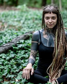 a woman with dreadlocks sitting on the ground surrounded by green plants and trees