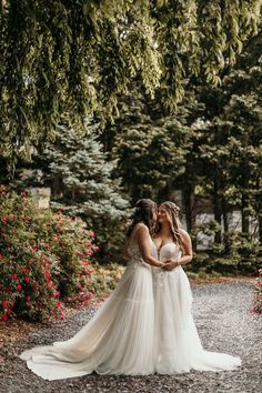 two brides standing in the middle of a gravel road surrounded by trees and flowers