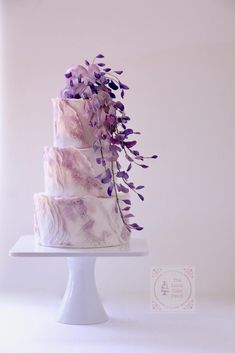 a three tiered cake with purple flowers on the top and bottom, sitting on a white pedestal