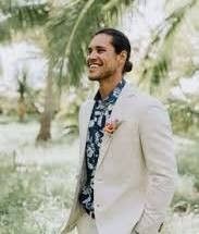 a man in a white suit and blue flowered shirt smiles as he walks through the grass