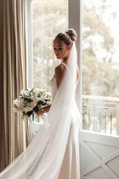 a woman in a wedding dress is standing by a window with her veil over her head