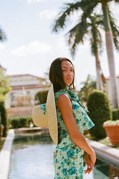 a woman standing in front of a pool wearing a green and white dress with a straw hat on her head