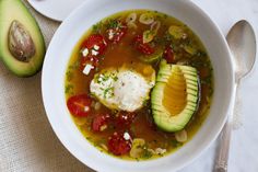 an avocado and tomato soup in a white bowl with spoons next to it