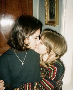 two women are kissing each other in front of a wooden door with a painting on the wall behind them