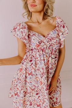 - You will look enchanting in this darling dress! - Lightweight material with a pink and purple hued floral print and a built-in lining - A v-cut neckline with gathered detail - Short flutter sleeves with an interior non-slip strip - A stretchy smocked back with zip closure - Hidden side pockets - A figure flattering silhouette that ends in a straight mini dress length hemline Pink Ruffle Sleeve Dress With Floral Print, Pink Floral Print Dress With Ruffle Sleeves, Pink Ruffled Dress With Surplice Neckline, Pink Ruffled Surplice Neckline Dress, Pink Surplice Neckline Dress With Ruffles, Pink Floral Print Dress With Butterfly Sleeves, Pink Floral Print Dress With Flutter Sleeves, Summer Floral Print Dress With Butterfly Sleeves, Feminine Cream Floral Dress With Ruffles