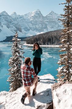 a man kneeling down next to a woman on top of a snow covered hill near a lake