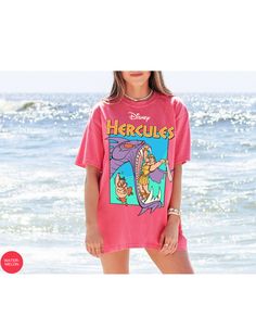 a woman standing in front of the ocean wearing a pink shirt that says hercules on it