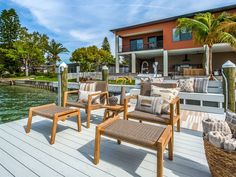 three chairs on a wooden deck next to the water and a building with palm trees in the background