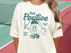 Introducing our Comfort Colors Pickleball Shirt "Dink Positive"- the perfect blend of comfort and style for the Pickleball player with a positive attitude! Crafted with the utmost care, this Unisex Pickleball Tee is made from premium Comfort Colors fabric, ensuring a soft, cozy feel that keeps you comfortable throughout your game. Whether you're hitting the courts for a fierce match or simply want to flaunt your love for Pickleball in style, this Pickleball Shirt is your go-to choice.  Made with 100% with ring-spun cotton, this soft-washed, garment-dyed fabric brings extra coziness to your wardrobe while the relaxed fit makes it an excellent daily choice. The double-needle stitching throughout the tee makes it highly durable while the lack of side-seams helps the shirt retain its tubular s Pickleball Tshirt Design, White Relaxed Fit Tennis Top, White Relaxed Fit Top For Tennis, Green Graphic Print Tennis Tops, Sporty Pre-shrunk Tennis Tops, Casual Tennis Tops With Screen Print, White Tennis Top With Team Name, Pickle Ball Shirt, Tshirts Design Ideas