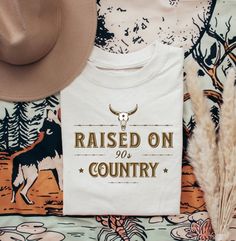 a t - shirt that reads raised on country with a cowboy hat and feathers next to it