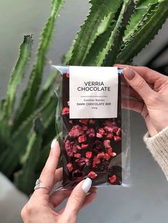 a person holding up a bag of chocolate next to a green plant with red flowers