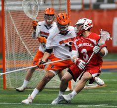 the men are playing lacrosse in orange and white uniforms with helmets on, while one man is running towards the goal